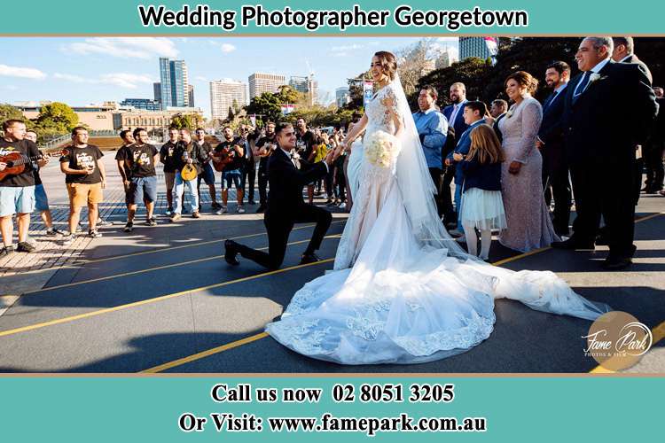 Photo of the Groom on his knee taking the hand of his Bride witnessing by the crowd Georgetown NSW 2298